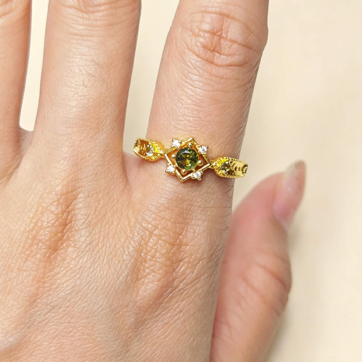 A gold ring with a central green gemstone and small clear accent stones, shown worn on a finger against a light beige background.